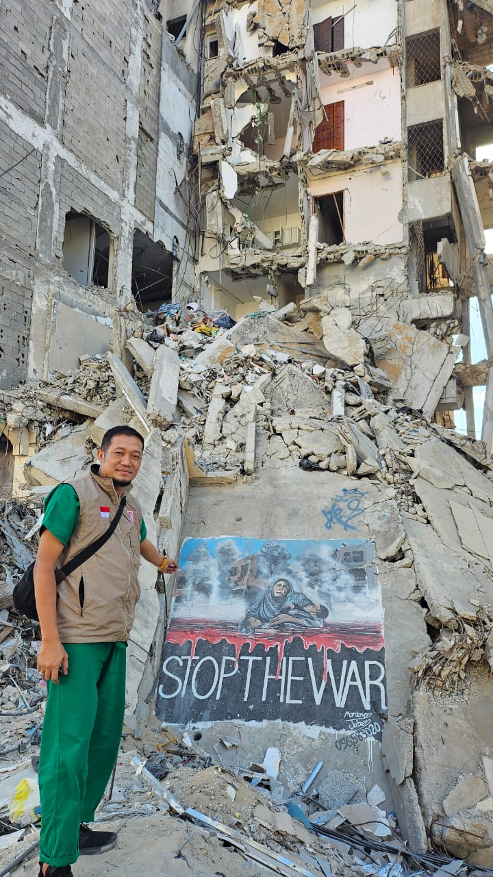 A person standing among rubble in Gaza beside a mural reading 'Stop The War'